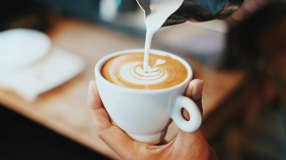 Kaffepaus En hand häller mjölk i en kopp kaffe med en konstnärlig latteart-design.